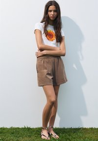 Camicia bianca a maniche corte con stampa di girasoli, pantaloni marroni e sandali piatti a fantasia. Modella in piedi sull'erba contro un muro bianco.