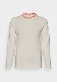 Béžová košile s dlouhým rukávem a kulatým výstřihem, která má kolem límce oranžový nápis "Calvin Klein Jeans", hladký přední díl a slim fit.