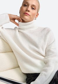 Witte gebreide coltrui met ribbeltextuur, relaxed fit en hoge kraag. Model draagt zilveren creolen en zwarte broek.