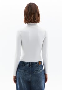 Langärmliges weißes Rollkragenoberteil, kombiniert mit hochgeschnittenen blauen Jeans. Verfügt über eine glatte Textur und eine lockere Passform, mit Gesäßtaschen.