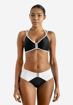 Woman wearing black and white two-piece swimwear with high-waisted bottoms and a strappy top, standing against a plain white background.
