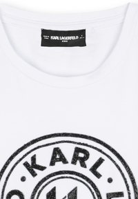 Camiseta de algodón blanca con un diseño de logo circular negro que incluye el nombre "KARL" y estrellas, cuello redondo y una textura suave.