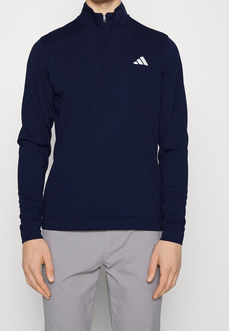 Pull-over à manches longues marine avec un zip quart, arborant un logo blanc sur la poitrine. Tissu doux et extensible avec des poignets côtelés.