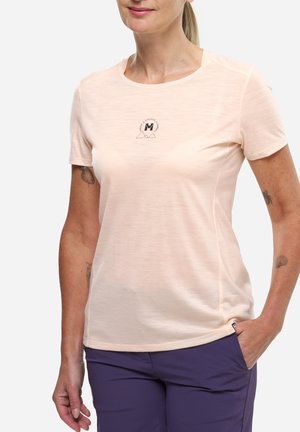 Femme portant un t-shirt rose clair à manches courtes avec un petit logo circulaire et un pantalon violet foncé, une main dans la poche, sur fond blanc.