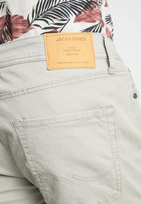 Pantalones gris claro con un parche de cuero etiquetado "JACK & JONES". Presentan un bolsillo trasero con un sutil detalle de costura y una textura suave.
