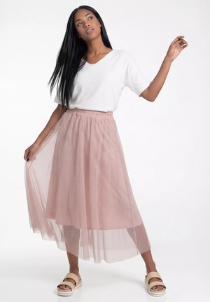 Kobieta z długimi czarnymi włosami, ubrana w biały top z krótkim rękawem, różową prześwitującą spódnicę midi i beżowe sandały, pozująca na białym tle.