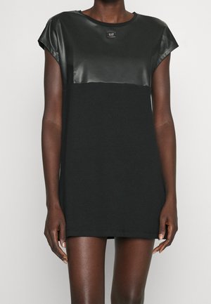 Robe noire à manches courtes avec panneau en similicuir et logo EA7 sur la poitrine, portée par une personne debout à la peau foncée.