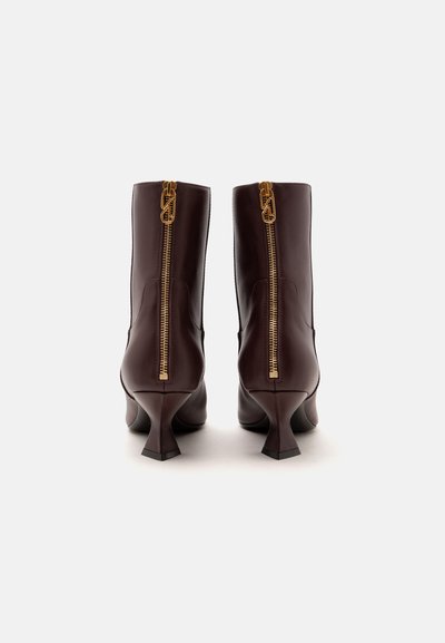Paire de bottines en cuir marron foncé avec fermetures éclair dorées au dos et talons hauts inclinés, vues de dos sur fond blanc.
