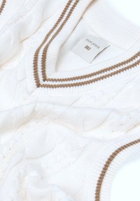 Chaleco de punto blanco con escote en V, patrón de trenzado texturizado y acentos a rayas marrones en el cuello y en las sisas. Etiqueta de la marca visible.
