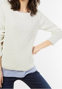 Vaalean kerma-nuori neule, jossa on teksturoitu neulekuvio, pyöreä pääntie ja kolme neljännestä hihoista, kerrostaen sinivalkoisen raidallisen paidan päällä.