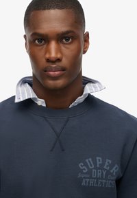 Donkere marineblauwe sweatshirt met een ronde hals en een lichtblauwe gestreepte kraag eronder. Bedrukt met de tekst "SUPER DRY ATHLETICS".