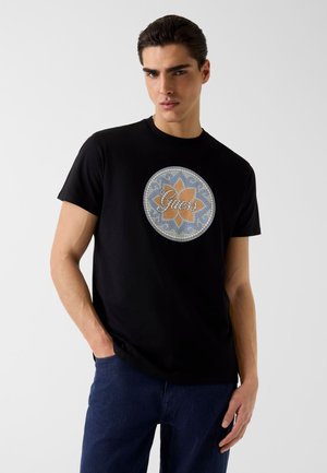 Jonge man met donker haar die een zwart T-shirt draagt met een cirkelvormig mozaïekontwerp en het woord "Guess", gecombineerd met donkerblauwe spijkerbroek.