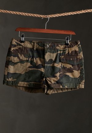 Cargo-shorts med camouflage-mønster hængende på en træbøjle fastgjort til et reb mod en mørk baggrund.