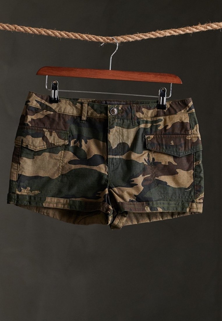 Short cargo à motif camouflage suspendu à un cintre en bois accroché à une corde sur un fond sombre.