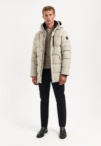 Lichtgrijze pufferjack met een capuchon, voorzien van een rits en elastische manchetten. Draagt over een beige trui en zwarte broek, gecombineerd met zwarte laarzen.