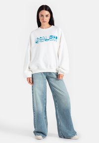 Biała bluza z niebieskim graficznym nadrukiem, w luźnym fasonie, z prążkowanym dekoltem i mankietami. Zestawiona z jasnoniebieskimi dżinsami o szerokich nogawkach.