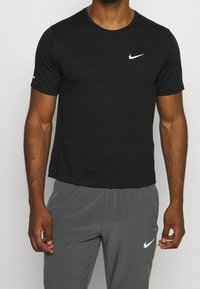 Camiseta atlética negra hecha de material ligero, con cuello redondo y logo de Nike en blanco en el pecho. Combinada con pantalones grises.