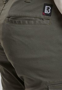 Olívové nohavice vyrobené z bavlnenej zmesi, s jednou zadnou kapsou a logom s geometrickým dizajnom v čiernej a marhuľovej farbe.