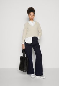 Beżowy sweter wykonany na drutach nałożony na białą koszulę, zestawiony z granatowymi szerokimi spodniami. Model trzyma czarną skórzaną torbę. Białe sneakersy.
