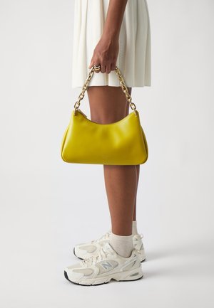Yellow leather handbag with a curved shape and gold chain handle, displayed against a light background, held by a person in a white dress.