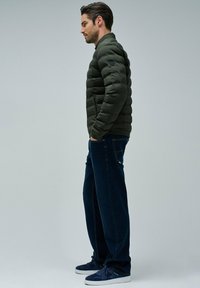 Hombre vestido con una chaqueta acolchada verde oscura, jeans azul oscuro y zapatillas de marino, de pie de perfil con las manos en los bolsillos frente a un fondo liso.