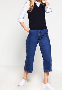 Námořnický bezrukávový svetr přes světle modrou košili s límečkem, v kombinaci s zkrácenými džínami. Model má na sobě bílé slip-on boty.