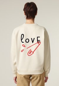Felpa oversize color avorio con ricamo del testo "love" in nero, provvista di un fermaglio di sicurezza rosso e gocce, maniche e orlo a costine.