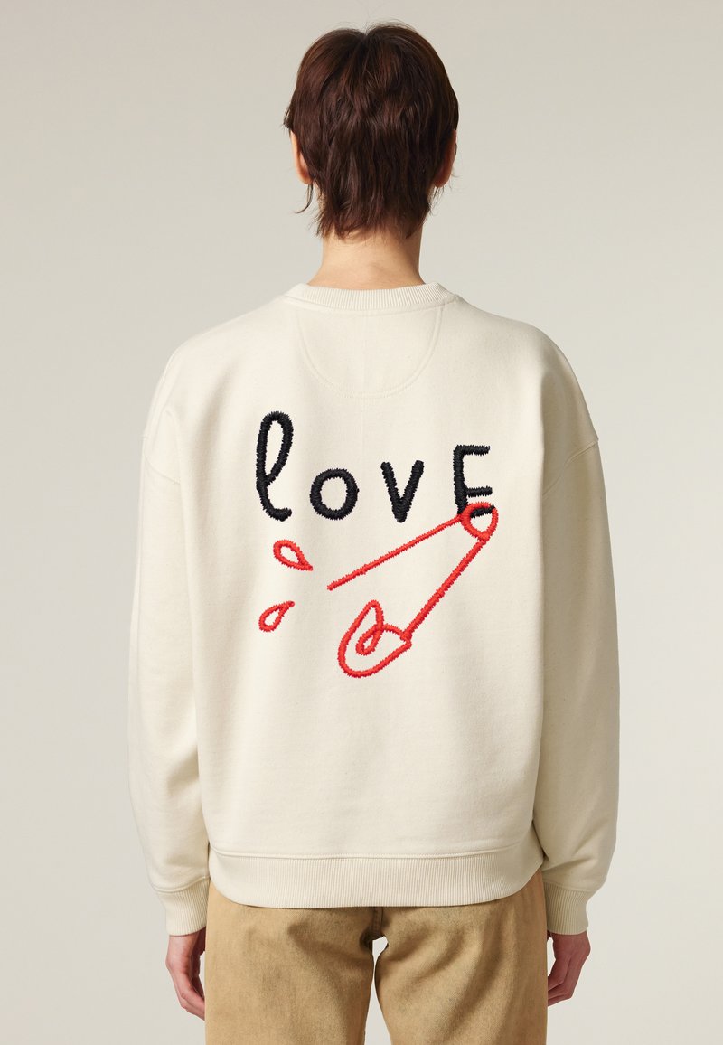 Felpa oversize color avorio con ricamo del testo "love" in nero, provvista di un fermaglio di sicurezza rosso e gocce, maniche e orlo a costine.