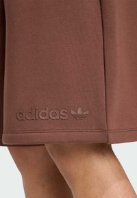 Hnedá mikina Adidas s textúrovanou tkaninou. Obsahuje embosované logo a emblém na leme. Priamy strih s akcentmi v podobe prešití.