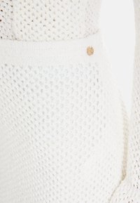 Weißer Strickpullover mit strukturiertem, offenem Gewebe, ausgestattet mit einem soliden Rippbund und einer kleinen kreisförmigen Logoverzierung.