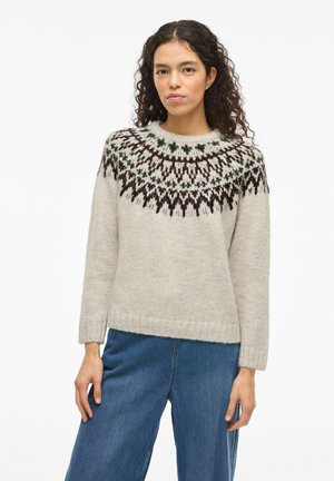 Femme aux cheveux bouclés et foncés portant un pull crème avec un empiècement à motifs noirs et verts, et un jean bleu ample, se tenant devant un fond blanc.