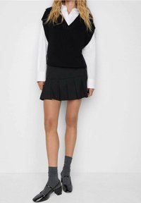 Schwarzer ärmelloser Pullover über einem weißen Kragenshirt, kombiniert mit einem plissierten schwarzen Rock, grauen kniehohen Socken und schwarzen Lackschuhen.