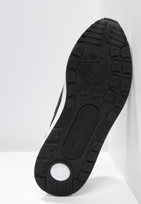 Svart gummisul med texturerat greppmönster, vita accenter och ett runt emblem. Har ett framträdande mönsterdesign för grepp.