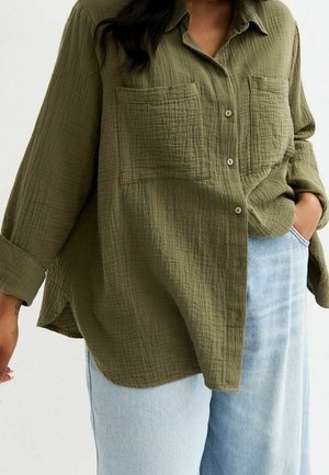 Persona que lleva una camisa suelta de color verde oliva con textura y botones, con dos bolsillos en el pecho, combinada con vaqueros azul claro, mano en el bolsillo.