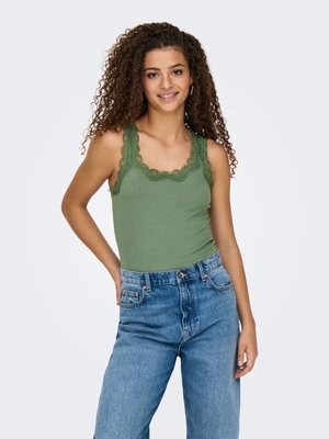 Jeune femme aux cheveux bouclés portant un débardeur vert avec finition en dentelle et un jean bleu taille haute, debout avec une main sur la hanche.