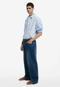 Helles blaues gemustertes Hemd mit hochgekrempelten Ärmeln, kombiniert mit lockeren dunkelblauen Jeans und hellbraunen Slippern.