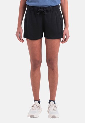 Person iført sorte shorts med snor, blå skjorte og hvide atletiksko, stående foran en ensfarvet hvid baggrund.