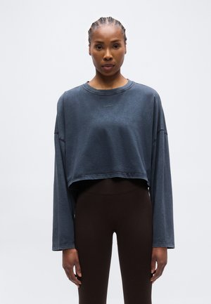 Kvinne med flette hår iført en løs, kort mørkegrå genser med lange ermer og tettsittende svarte leggings, stående mot en ensfarget lys bakgrunn.