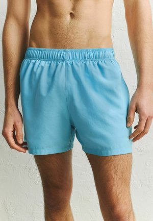 Mężczyzna ubrany w jasnoniebieskie spodenki z elastycznym pasem, stojący na tle teksturowanej białej ściany, pokazujący tors i górną część nóg.