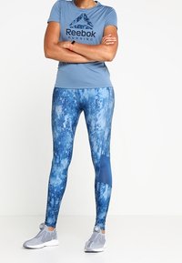 Maglietta da corsa Reebok di colore azzurro con logo, abbinata a leggings blu con motivo e scarpe da ginnastica grigie con un design testurizzato.