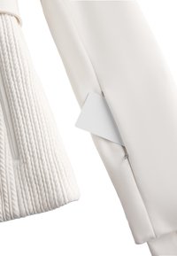 Giacca bianca con manica a coste testurizzata, tessuto liscio e una tasca con zip che mostra un piccolo'etichetta bianca. Design pulito e minimalista.