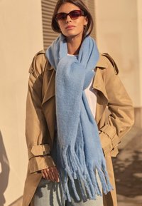 Heledsinine, pehme villane sall frantsi lõpudega, kantud beežist trench coat'ist. Mudel seisab helehallil seinal, kandes prille.