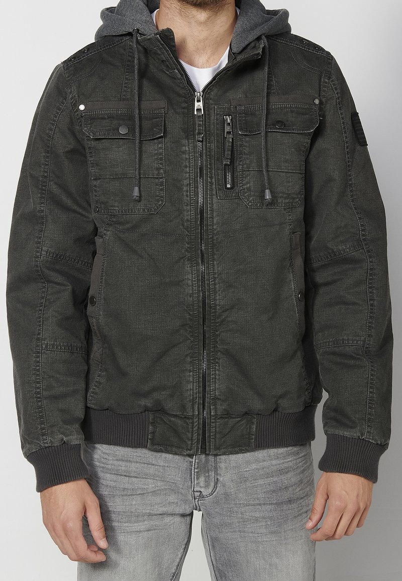 Homme portant une veste zippée vert foncé avec poches poitrine boutonnées et capuche grise attachée, associée à un jean gris clair.