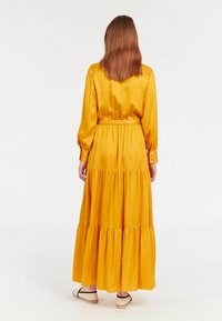 Robe satin jaune longue avec des manches longues, taille élastique et ourlet à volants en niveaux. Boutons aux poignets des manches ; texture lisse et brillante.