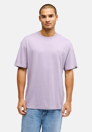 Jeune homme portant un t-shirt oversize violet clair uni et un jean bleu, debout devant un fond blanc uni.