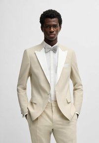Tuxedo beige con rever in satin bianco, abbinato a un papillon argentato e una camicia bianca impeccabile; tessuto morbido, vestibilità su misura e dettagli della tasca.