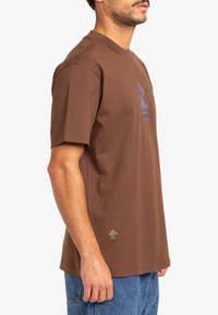 Brown cotton t-shirt with short sleeves, featuring a blue graphic print on the chest and a subtle logo accent near the hem.