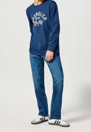 Camiseta de manga larga azul marina con el gráfico blanco "Wrangler 1947" y banderas a cuadros. Combinada con unos vaqueros de mezclilla azules y zapatillas negras y blancas.