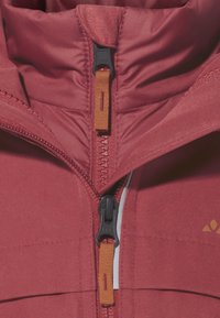 Gros plan sur l'avant d'une veste isolée rouge avec deux zips noirs, des pulls en tissu orange et un petit logo partiellement visible sur le côté droit.