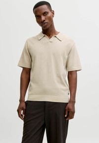 Pull polo beige à manches courtes en maille avec un col plat, présentant une finition texturée et un ourlet côtelé. Porté avec un pantalon foncé.